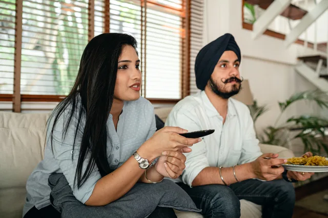 Calm Indian woman with remote control and man in turban and with mustache having meal while watching TV on couch. Connected TV advertising: planning a local campaign