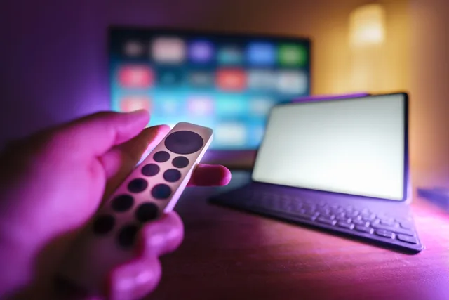 A hand using a remote to control a TV and laptop in a cozy room setting, perfect for relaxation. A connected TV advertising market entry checklist for English buyers