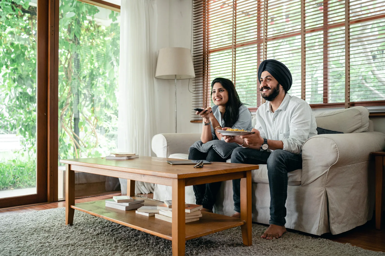Cheerful Indian woman and man in turban spending free time together while watching television and sitting on sofa in living room near table
