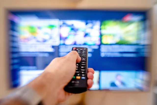 Hand holding remote control pointing at a blurred TV screen in a cozy indoor setting. Connected TV advertising dashboards: metrics and governance