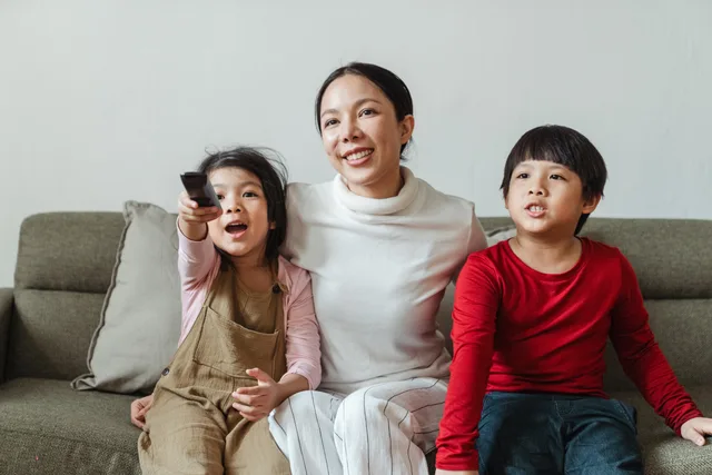 Optimistic Asian woman embracing little daughter and son in casual outfit sitting on sofa while choosing TV channel together using remote. Why a connected TV advertising selection checklist prevents rework