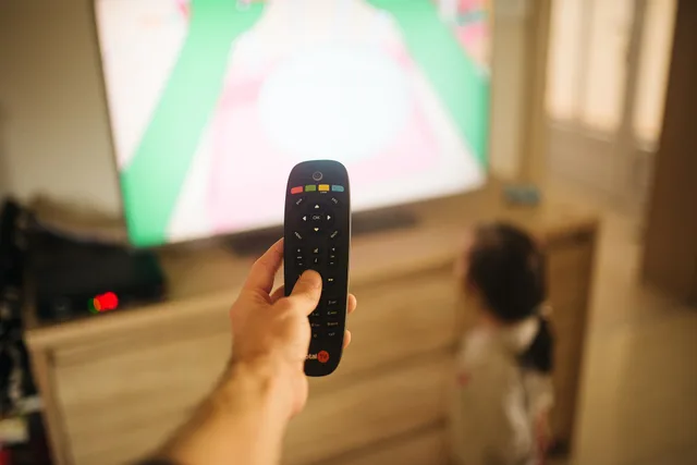 A child sits on the floor and watches a colorful television show. An adult holds a remote control aimed at the TV. Sunlight comes in through the window. Sky AdSmart vs ITVX: which platform fits your CTV campaign?